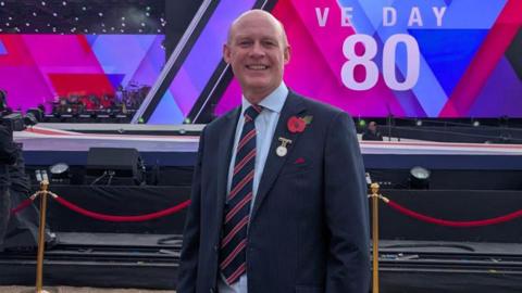 A man stands in front of a stage with a red, pink and blue backdrop and the words "VE Day 80" written in white. He has a shaved head and he is wearing a dark blue suit. He has a small gold medal and and poppy pinned to his jacket.