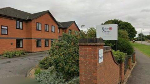 A brown two-storey building is located within a small driveway leading up to it. At the front is a fence with a white sign which reads Kesteven Grange, surrounding by shrubbery. A path and road can be seen in front of it.