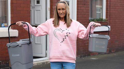 Tracy Harries stands in a street of red brick terraced houses with white upvc doors and windows. She is wearing a pink sweatshirt with a heart emblem and pale jeans and has long straight fair hair and is holding two grey food waste collection caddies out to each side
