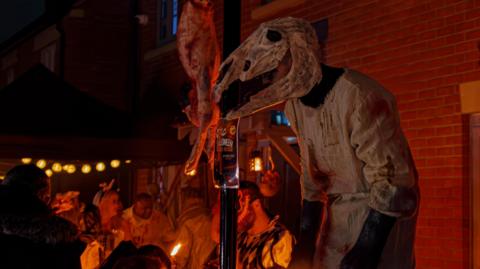 A person wearing a horse head skeleton costume and dressed in a bloody white robe stands beside the entrance to the Brackley Horror House 