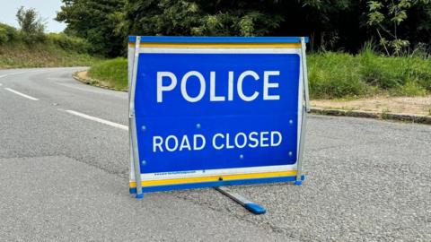 A blue metal sign standing on a tarmac road that reads POLICE ROAD CLOSED. There is a grassy verge and trees along the road.