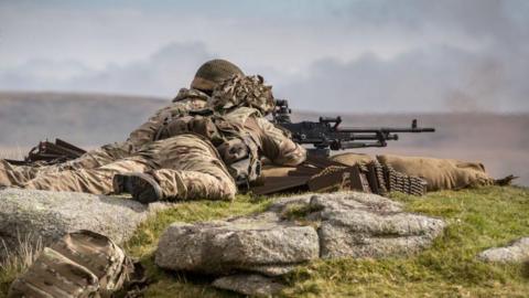 Soldiers from 6 RIFLES fire live ammunition from a General Purpose Machine Gun on the range at Okehampton Camp, Dartmoor, during a training exercise in 2017. They are wearing camouflage and lying on the ground as they load a machine gun