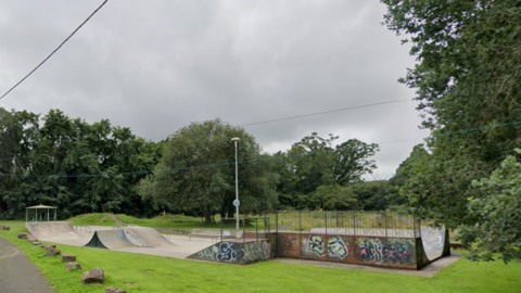 Skatepark in Okehampton. Graffiti is on the side of the skatepark. Grass is surrounding the site. 