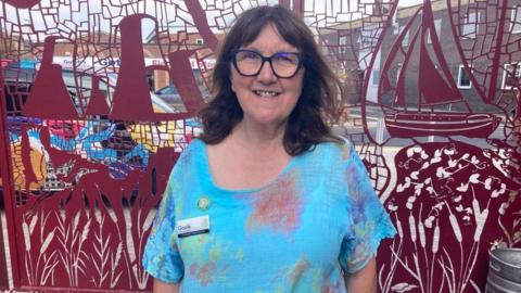 A woman with shoulder-length brown hair and wearing blue-rimmed glasses is looking into the camera and smiling. She is standing in front of a burgundy-coloured metal structure and wearing a bright blue floral blouse.