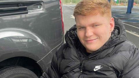 Ralph Pearce has short ginger hair and is wearing a black coat while sitting in a wheelchair in a car park. He is smiling.