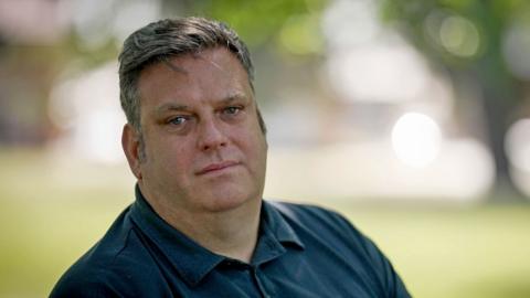 Steven is wearing a blue shirt and is sitting on a bench outside. He is in focus but the background, which resembles grass and trees, is fully out of focus. Steven is looking directly into the camera with a straight face. 