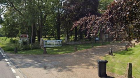 The driveway to Deene Park, near Corby, showing a brown heritage sign saying Deene Park and a colourful picture of the stately home