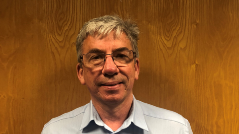 A man in glasses and with grey hair looking straight on. He is in a smart blue shirt and is stood in front of a wooden panel