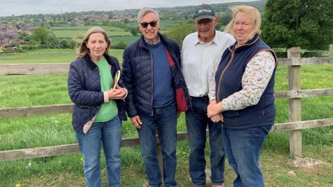 Two men and two women are dress casually in jeans and outdoor jackets and are standing by a fence overlooking a field