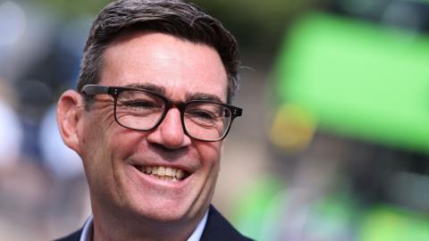 Andy Burnham is smiling and looking to the side. He has dark hair, brushed to one side and dark glasses.