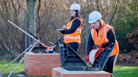Two women in orange hi-vis jackets and white hard hats working at a site. The woman on the right has blonde hair and is holding some wire and unwinding it into what looks like a brick chimney on the ground. The woman on the left is slightly further back and has long brown hair. She is holding a laptop in her hand. 