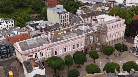 A large pink building from above, with trees at the front and other buildings behind it 