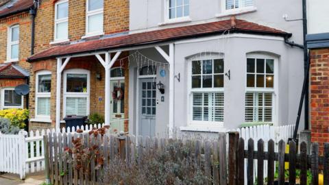 A row of terraced houses with brick and painted facades, white-framed windows, small front gardens, and low wooden fences along a residential street.