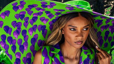 Models wait backstage wearing huge hats and clothing featuring the same green-and-purple print.