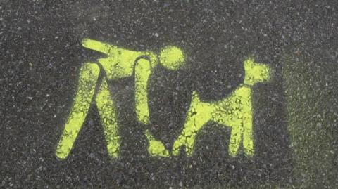 A yellow sign painted on a pavement shows a person cleaning up after their dog