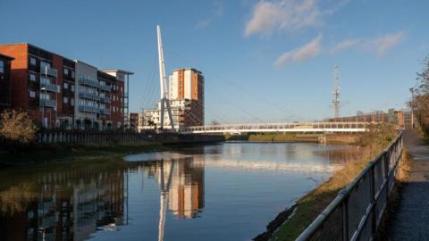The cable-stayed 'Sir Bobby Robson' pedestrian footbridge over the River Gipping, Ipswich on a sunny day. Large blocks of flats line the left hand side of the river while there is a footpath running on the right hand side. 