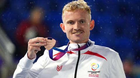 Mullarkey stood, holding his bronze medal up. His jacket is an Isle of Man team jacket.