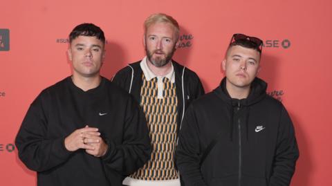 A picture of the members of the group. They  are looking at the camera while standing in front of a red background. 