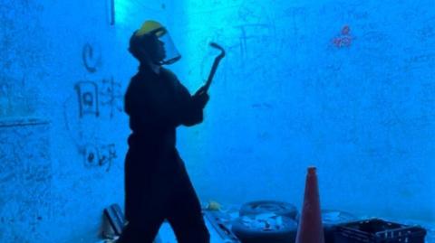 Picture of someone in a blue light-lit rage room surrounded by items to smash. They are wearing a yellow helmet and holding a crowbar. Among the items are a crate, car tyres and a traffic cone. There is rudimentary graffiti on the walls.