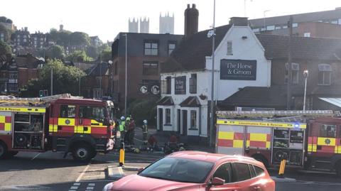 A street showing two red fire engines from Lincolnshire Fire & Rescue, with firefighters attending to an incident at a white two-storey pub with a sign reading The Horse & Groom. A red Ford Focus car is in the foreground. In the background, a steep hill rises, dotted with houses, and the three towers of a cathedral can be seen rising above other buildings.