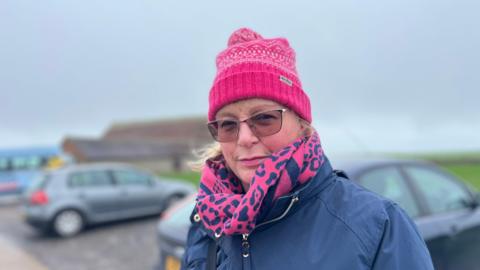 Emma Dyer a seaford resident standing in the car park with the barn and cars behind her. she's wearing glasses, a pink scarf and pink hat