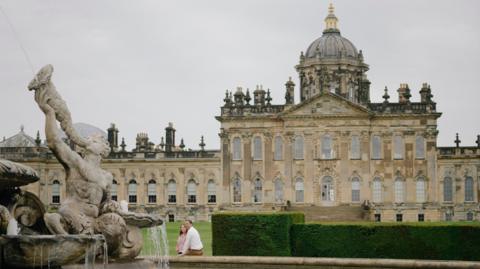 Castle Howard. A large stately home. A couple sit on the edge of a fountain in front of the property. 