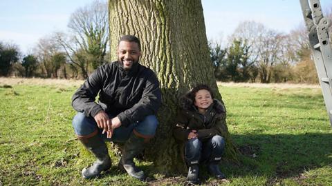 JB Gill leans on a tree with his son Ace - both are wearing cold weather clothing and wellies. 