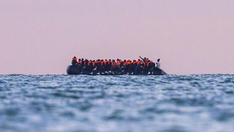 A side view of a small rubber boat filled with people wearing life jackets crosses the English Channel towards the UK.