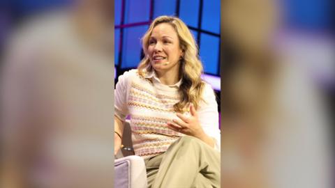 Eline Van der Velden has long blonde hair. She is seated, and wearing a white shirt, sleeveless jumper and beige trousers.