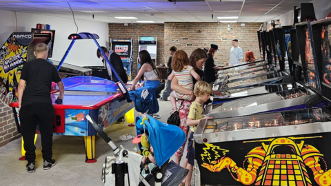 Inside the arcade with several brightly coloured pinball machines lined up against one wall and people, including children, playing on them. There is also an air hockey table with young people playing.