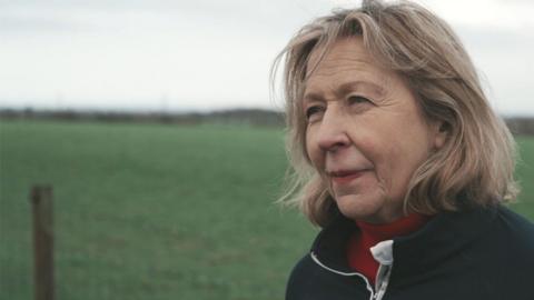 Chris Pollitt, with shoulder-length blonde hair, wears a red polo-necked jumper and a black cardigan as she stands in front of a green field.