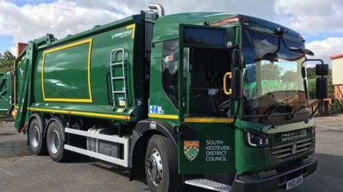 A photo of a forest green bin lorry with bright yellow markings. It is parked diagonally with the front of the lorry facing the camera. The driver's door is open and has a graphic that reads "South Kesteven District Council" and has the authority's logo next to it. 