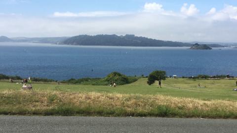 A view from a car park across a green cliff in front of blue sea and showing an island and Cornwall in the distance