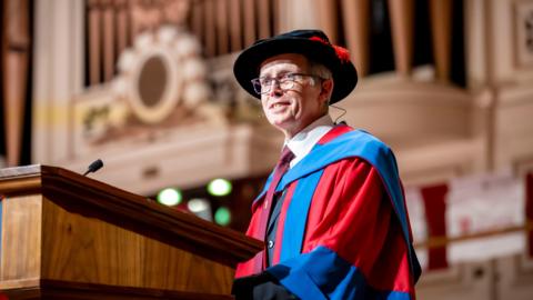 Dr Nigel Hewett wearing a red and blue gown, black hat. He is standing at a wooden podium that has a microphone on top.