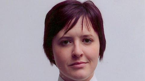 Philippa Reynolds in a police photo. She has short purple/red hair and is wearing her uniform. She is standing in front of a white wall. 
