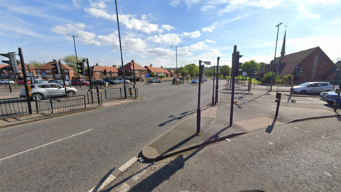 A large cross roads, dotted with traffic lights and crossing points. The road is surrounded by residential semi-detached houses and a church can be seen on the left.