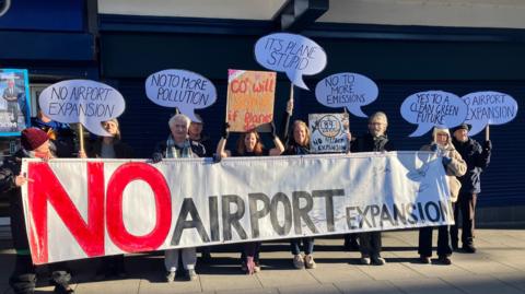 A dozen campaigners with placards stand behind a white sign which says "No Airport Expansion". Behind them is a shop with its shutters down. 