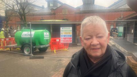 Photograph of shopkeeper Val Unwin, who runs a store in the centre of Ashton-under-Lyne, during the redevelopment work. Part of the background is fenced of and workmen wearing yellow hi-vis clothing are at work.