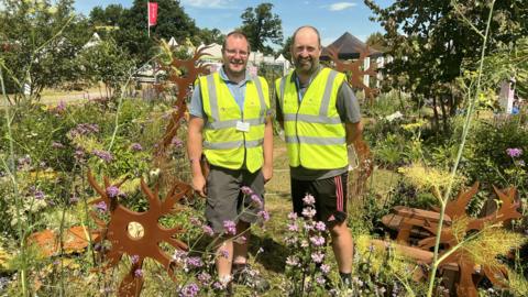 Two men wearing hi-vis stand in the garden they've created with many flowers and sculptures at the Badminton Flower Show