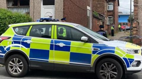 A police car in front of cordoned-off alleyway with police officers visible behind it.