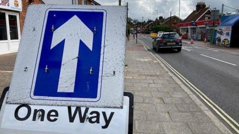 A blue one way sign on the pavement next to a road lined with shops