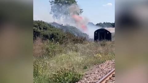 Smoke and flames can be seen from the side of a train, surrounded by vegetation. It is a bright and clear day.