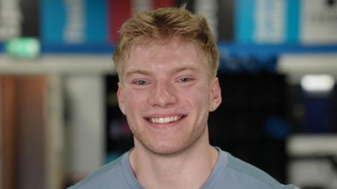 Close-up of a person in a light blue sports top, facing the camera and smiling. The background is softly blurred, showing an indoor gym.