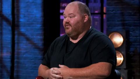 A close up shot of Simon Sansome sitting in front of the dragons pitching his app. He is wearing a black t shirt.