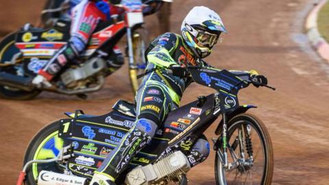 A speedway racer steers his bike around a corner on a mud track. Other racers can be seen behind him.