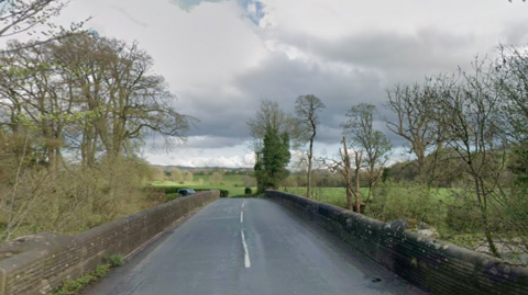 A two-lane bridge runs over a river, with trees and countryside all around.