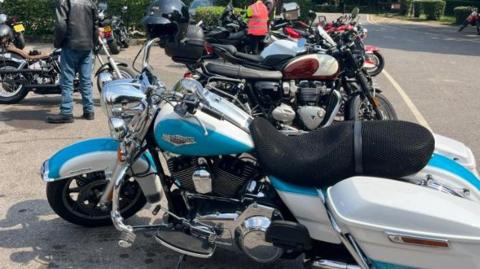 Various motorbikes queued up at a carpark in Micklesham, the font bike is blue and white with a dark coloured seat. 