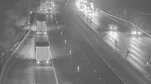 Cones can be seen on a black and white image of the motorway closing off some lanes. Traffic is driving on the remaining lanes.