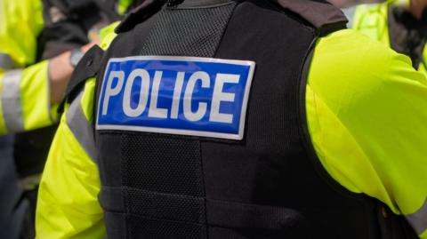 Close-up of 'POLICE' marking written on the back of a hi-visibility stab proof vest worn by a trio of police officers at the scene of an incident. 