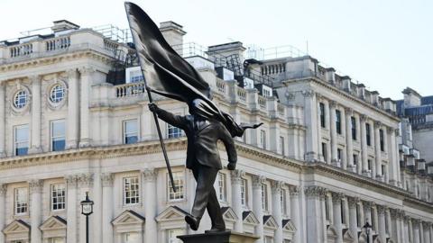 the statue of a man in a suit walking off a plinth holding a flag that is covering his face.
